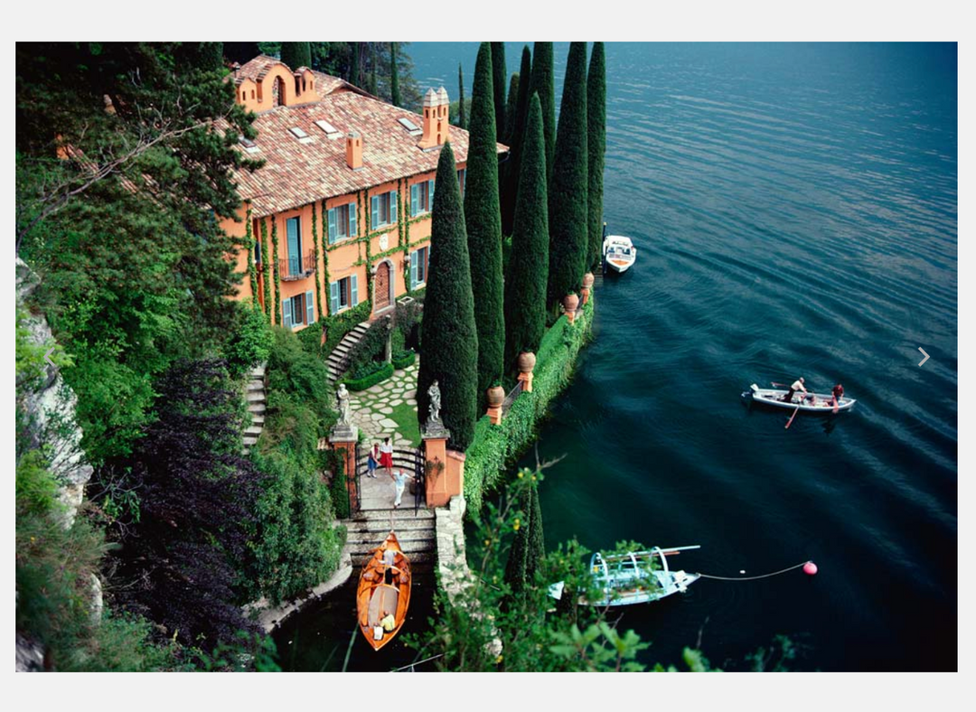 A scenic photograph of a large orange villa on a hillside beside a deep blue lake, with tall dark cypress trees, a stone staircase, boats on the water, and a white border around the image. Lake Como